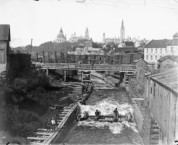 A black-and-white photograph of two people on a lumber raft down the Chaudière timber slide, with Parliament buildings in the background.