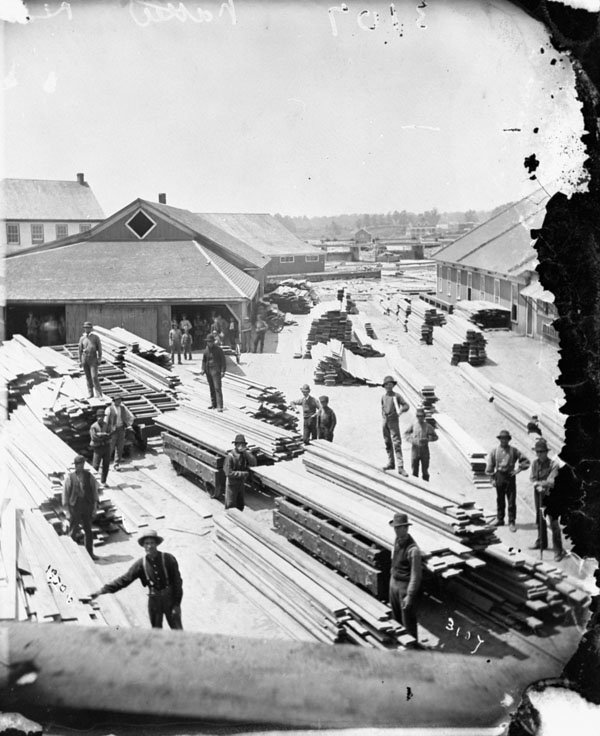 A black-and-white photograph of a lumberyard, with several men at work among piles of lumber in the foreground, and mill buildings in the background.